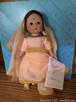 Front view of Madame Alexander India doll, dressed in peach traditional outfit with gold trim and sheer dupatta, holding original tags, sitting in original blue box with pink Madame Alexander print.