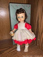 Madam Alexander doll standing in front of original box, wearing red dress, white apron, and holding basket with white duck