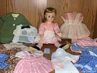 Madame Alexander doll seated among several clothing items including coats and dresses, showing the quantity and variety of garments included.
