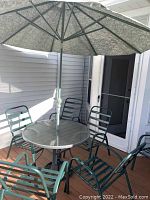 Full view of round grey metal patio table with attached sage green patterned umbrella and six dark green metal chairs around the table on wooden deck.