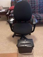 Black office chair with cushioned seat, backrest, armrests and five-wheel base. Black paper shredder placed on the floor beside chair.