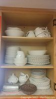 Open cabinet showing stacks of white ceramic dinner plates, salad plates, dessert plates, bowls, teapot, cream and sugar set, and serving platter on multiple shelves.