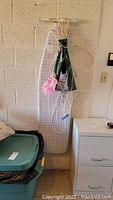 Ironing board hanging against wall with apron, clothesline, and bag hanging on it.