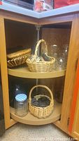 Corner cabinet shelves with several clear glass canning jars, including jars with lids and jars without lids. Also visible are multiple woven wicker baskets, some with handles, arranged on two shelves.