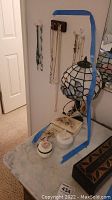 Photo showing several costume jewelry necklaces hanging on wall hooks above three small trinket boxes on a marble surface next to a Tiffany-style lamp.
