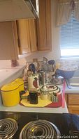 Photo showing yellow lidded containers, apple-themed utensil holder, various cooking utensils, and tea kettle on kitchen counter.