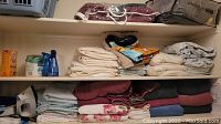 Three shelves showing stacks of folded towels, sheets, linens, a clothes iron, and household cleaning items.