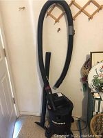 Full view of black Craftsman shop vacuum with hose attached, hanging on a wall and standing against a corner.