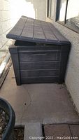 Closed large dark grey plastic outdoor storage tote beside a house wall.