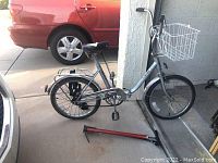 Side view of the silver folding bicycle with black seat and white front basket, tires flat. Red bicycle hand pump lying on the floor beside bike.