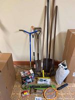Photo of various garden tools including a blue-handled soil rake, two shovels with wood handles, green garden hose, yellow bug zapper, boxed paint sprayer and gardening accessories on floor.