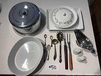 Photo showing covered silver serving dish, white oval ceramic serving bowl, ceramic butter dish with lid, silver teaspoons, pasta fork, large spoon, salt and pepper shakers, and metal scoop arranged on table.