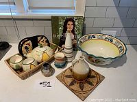 Photo showing the full kitchenware lot including the tea pot set in basket, large bowl, trivet with jug, tall vase, small pot, and cookbook