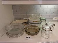 Photo showing Corning Ware baking dishes stacked with lids, glass jars with clamp lids, metal skewers, and a clear glass baking dish and serving bowl on wire rack.