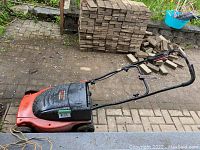Side view of Lawn Hog electric lawn mower showing orange base and black handle, with some dirt and a brick wall background.