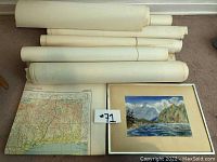 Lot overview showing the framed watercolor painting laid flat and multiple rolled aeronautical charts on the floor.