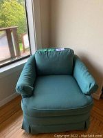 Front view of the green upholstered armchair in a corner by window with natural light. Shows the full chair with armrests and cushion details.