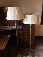 Two brass floor lamps side by side; taller lamp with a shade and a glass globe, shorter lamp with a beige pleated shade.