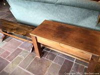 Photo showing two wooden primitive benches next to each other on tiled floor against blue couch. Low knotty pine bench with visible wood knots, and a taller storage bench in finished wood.