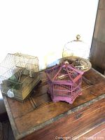 Image showing three vintage bird cages on a wooden surface. One wooden cage in the center with a pagoda structure, two brass cages on either side.