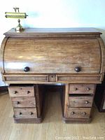 Front view of roll top desk with closed top and two rows of drawer fronts, two wooden knobs on roll top, brass lamp sitting on top.
