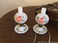 Two vintage white glass globe lamps with pink floral design on a dark wood table, shown unlit