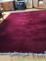 Full rug spread on wooden floor showing deep burgundy color and cream-colored fringe ends on one side and the surrounding environment including furniture.