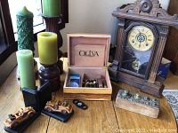 Wooden cigar box labeled 'Oliva' open with cigars and cigar tools inside, surrounded by four green candles on wood pedestals, two wooden car bookends, a block with metal weights, and a mantle clock.