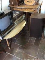 United Audio turntable positioned on wooden table with glass lid closed; adjacent to small Mitsubishi TV and one SRL floor speaker; Zenith antenna behind TV.