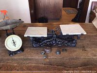 Full view of two antique scales on a wooden table, one a marble top balance scale with weights, the other a grocer dial scale.