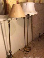 Two brass floor lamps standing side by side showing moveable arm on one and lampshades on both.