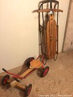 Full view of the vintage sled and kids row cart collected together viewed from side angle on plywood floor.