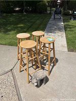 Four wooden bar stools arranged in a group outdoors on pavement with green grass and a wheelchair in background. A small carved bear-shaped plant stand is positioned near the stools.