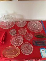 Glass cake stand with dome lid along with six glass bowls and four glass platters arranged on a red cloth.