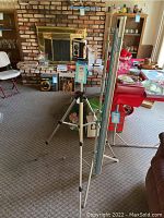 Brownie Reflex camera mounted on a silver tripod with a projection screen beside it in a living room.