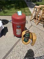 Red Charbroil smoker with a gray metal access door, next to Coleman lantern in a yellow carrying case on the ground outdoors.