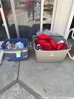Wide view showing two plastic storage bins, one blue and one beige, placed outdoors on concrete, containing holiday plush toys, decorations, and assorted smaller items.