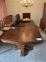 Front view of rectangular wooden dining table with a polished surface and carved pedestal legs, in a dining room setting with chairs and chandelier.