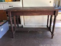 Full view of rectangular wooden dining table showing turned legs and stretcher base, placed on concrete floor against wall.