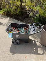Black plastic garden cart filled with tangled assorted extension cords, visible scuffs on cart surface, displayed outside on concrete near shrubbery.