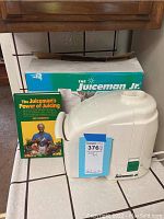 White Juiceman Jr. juicer on counter with original teal and white box and green-covered juicing book by Jay Kordich in front.