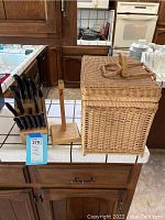 Photo of a kitchen cutlery block with black-handled knives, a wooden banana hook stand, and a wicker picnic basket on a kitchen counter.