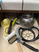 Drawer showing a white apple cutter with black handles, yellow juicer, metal steamer insert, black handled tongs, metal ice cream scoop, nutcracker, and measuring cups.