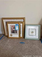 Four framed art pieces grouped on carpet, three with gold-tone frames, one with a blue-green wooden frame. Visible tags and seal on Emmett Kelly item.