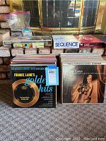 Photo of LP albums in cardboard sleeves stacked in two rows in front of a fireplace, showing album covers from Frankie Laine, Saint Brass, Eddie Peabody and others. Also visible are boxed board games Sequence and Uno on the hearth.