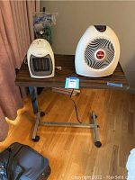 Photo of the two portable heaters placed on the adjustable bed table showing the Vornado heater on the right and Pelonis heater on the left with power cords visible.