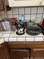 Kitchen countertop showing assortment of pots, pans, kettle, boxed dumpling press, and defrosting tray.