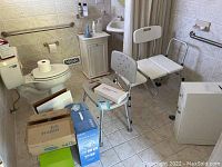 Wide view of bathroom featuring two shower chairs, various boxes including Homedics foot spa, raised toilet seat, and other items