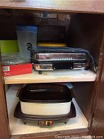 Wooden shelves featuring a chrome electric press/pasta machine on the top shelf and a West Bend slow cooker with ceramic insert on the bottom shelf, power cords visible.