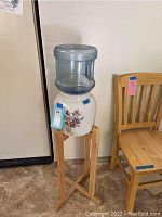 Side view of water jug showing clear plastic container on ceramic base and wooden stand with adjacent wooden chair.